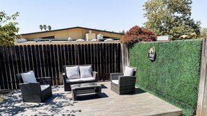Een terras buiten met rieten meubels, een glazen tafel en een groene haag.