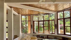 A spacious living room with a large beige sofa, a glass coffee table, and a ceiling fan. Large windows provide a view of lush greenery outside. - Tamarindo Grand Villa. Oasis at walking distance to Tamarindo Beach! (Tamarindo)