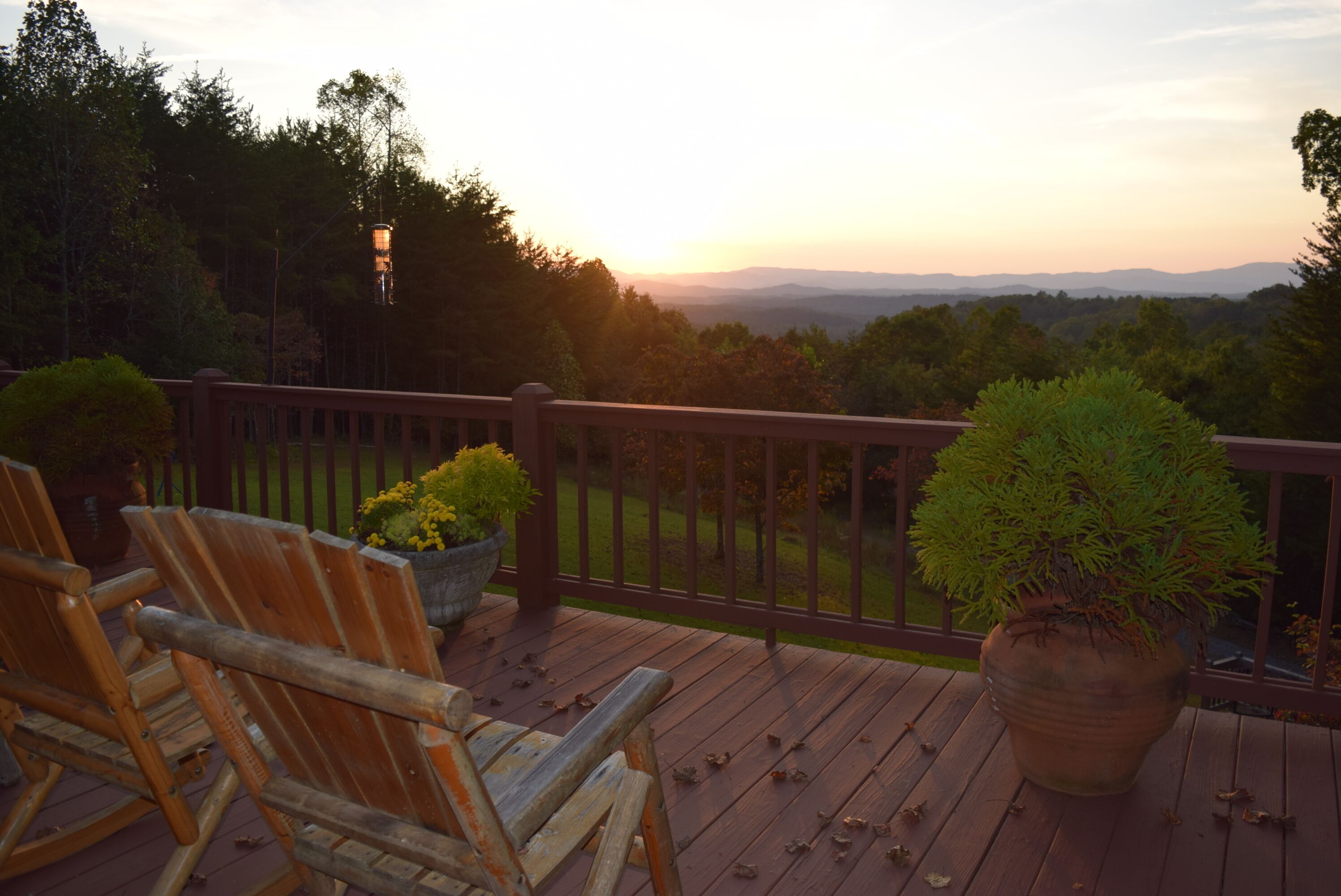 A wooden deck with rocking chairs and potted plants, overlooking a forested landscape.