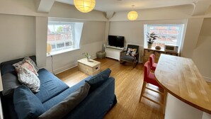Un salon moderne avec un canapé, un téléviseur, une table à manger en bois et deux fenêtres.