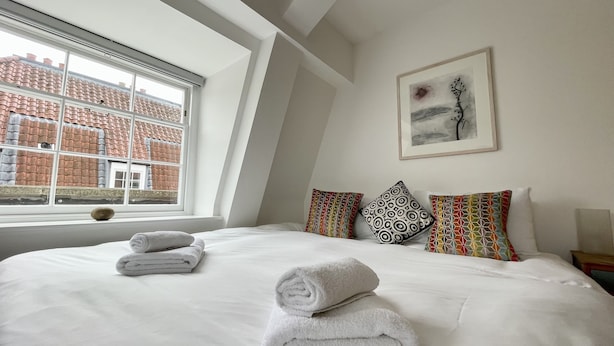 Une chambre avec un grand lit, du linge de lit blanc, une fenêtre donnant sur des bâtiments aux toits rouges et une œuvre d’art encadrée accrochée au mur.