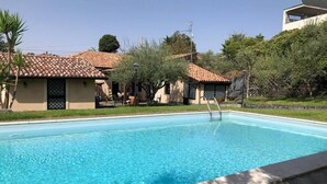 Una piscina con superficie d'acqua cristallina e blu, circondata da un solarium in pietra e una piccola area giardino con alberi e arbusti.