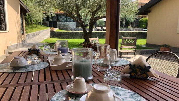 Tavolo di legno apparecchiato per la colazione, con tazze da tè, un boccale di latte e una brocca, all'aperto con vista su un giardino e un edificio.
