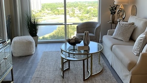 Un salon moderne avec une table basse en verre, un canapé blanc et une vue sur un quartier résidentiel grâce à de grandes fenêtres.