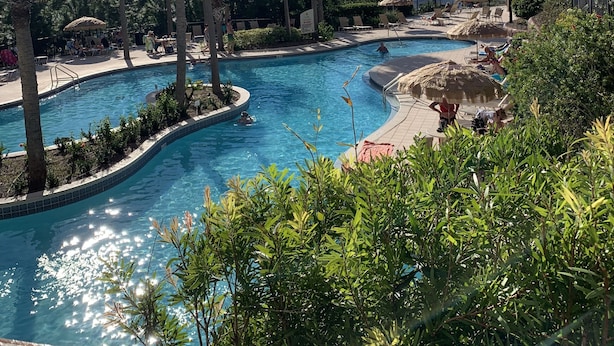 Un espace piscine aménagé dans un complexe hôtelier, avec des palmiers, des chaises longues et un ciel d’un bleu limpide.
