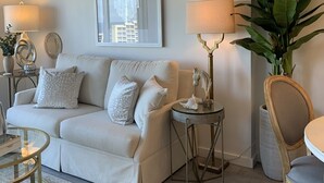 Un salon avec un canapé beige, deux tables d’appoint avec des lampes, une grande plante en pot et un tableau encadré au mur.