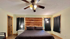 A bedroom with a bed, a nightstand, a chair, a ceiling fan, and two windows with curtains. - Poco's Retreat                                . (Many)