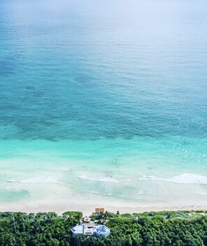 Vista aérea de una zona costera con mar azul cristalino, una playa de arena y un pequeño edificio rodeado de vegetación densa y verde.