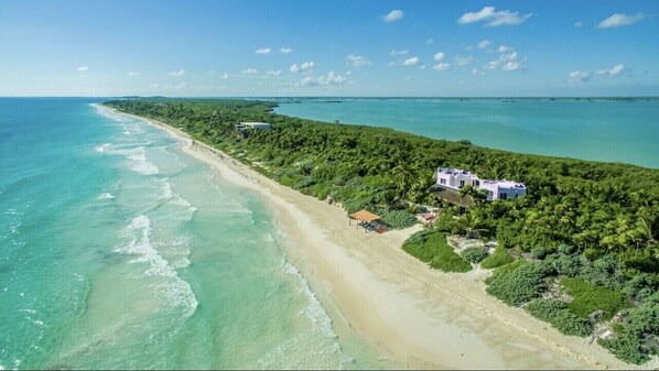 Luchtbeeld van een kustbezit met een strand, dichte begroeiing en enkele gebouwen.