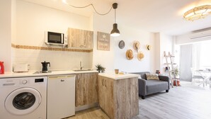 Une cuisine compacte équipée d’un lave-linge, d’un four à micro-ondes et d’une machine à café. Le salon comprend un canapé gris et une table à manger avec des chaises.