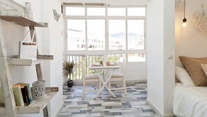 Une pièce dotée d’une grande fenêtre, d’un lit blanc et d’une table avec des chaises. On y trouve également des étagères remplies de livres et d’objets décoratifs.