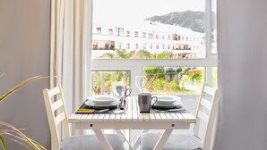 Une table à manger est dressée avec des assiettes, des verres et une tasse, placée près d’une fenêtre d’où l’on peut admirer un bâtiment et de la verdure.