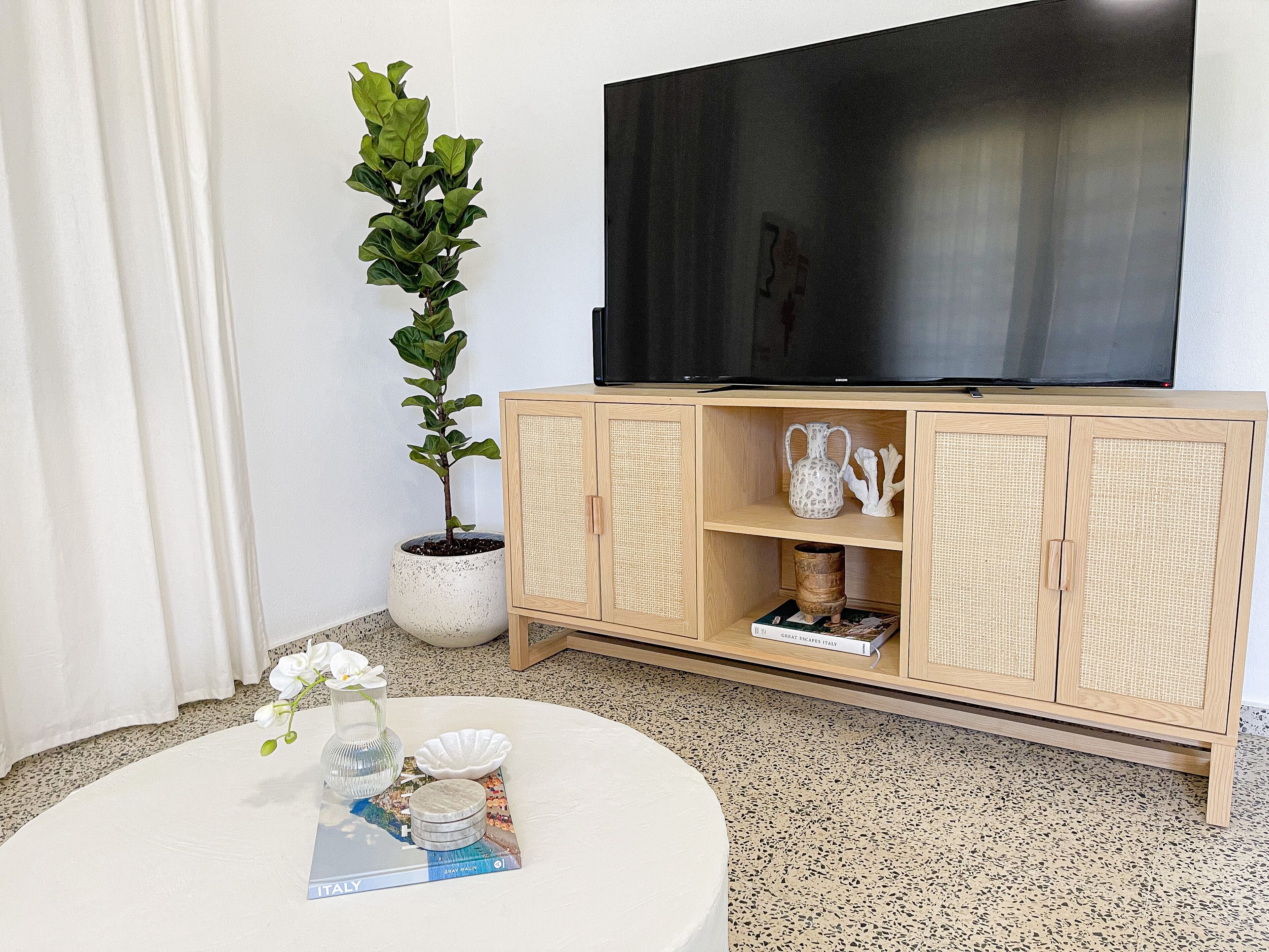 Sala de estar con televisor sobre un mueble de madera, una planta en maceta y una mesa de centro redonda con libros y objetos decorativos.
