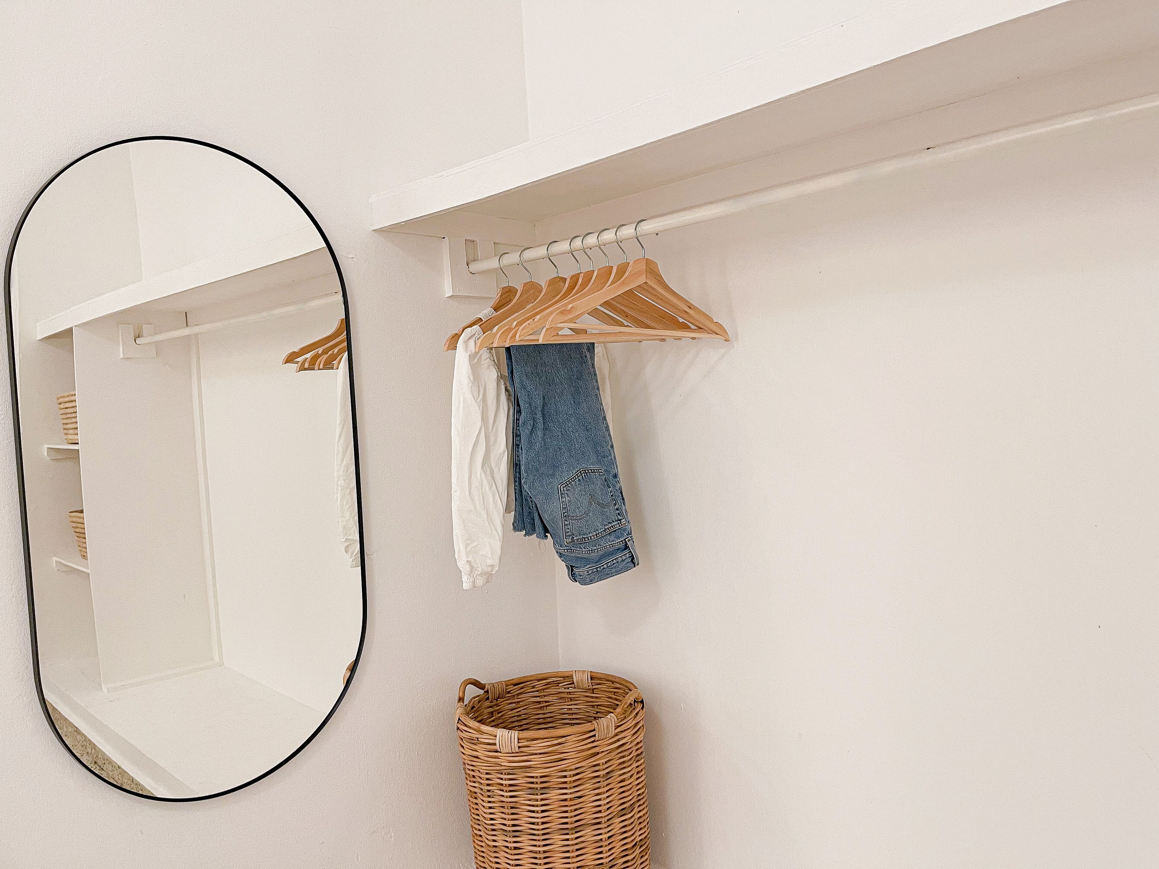 A white wall with a round mirror, a wooden hanger rack with clothes, and a wicker basket.