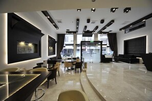 A modern hotel lobby with a central seating area, a reception desk, and large windows offering a view of the street. - Europa Hotel (Kastoria)