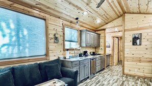 Interior de una cabaña de madera con sofá, cocina y un ventanal grande.