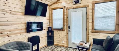 Interior de una cabaña con televisor, asientos, una puerta con ventanal y techo de madera.