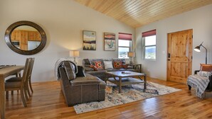 Un salon chaleureux avec un plafond en bois, un miroir rond, un canapé agrémenté de coussins colorés, une table basse et un coin repas avec une table et des chaises en bois.