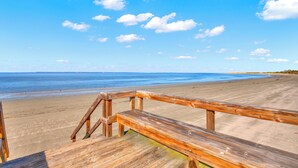 Une promenade en bois mène à une plage de sable fin, sous un ciel bleu clair parsemé de quelques nuages.