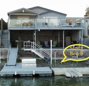 Una propiedad frente al agua con muelle, terraza cubierta y escalera que conduce a un segundo nivel con mobiliario exterior.