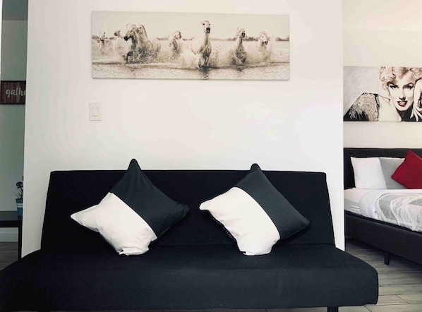 A modern room with a black sofa, two black and white pillows, a bed with white and red pillows, and a wall-mounted artwork of horses.