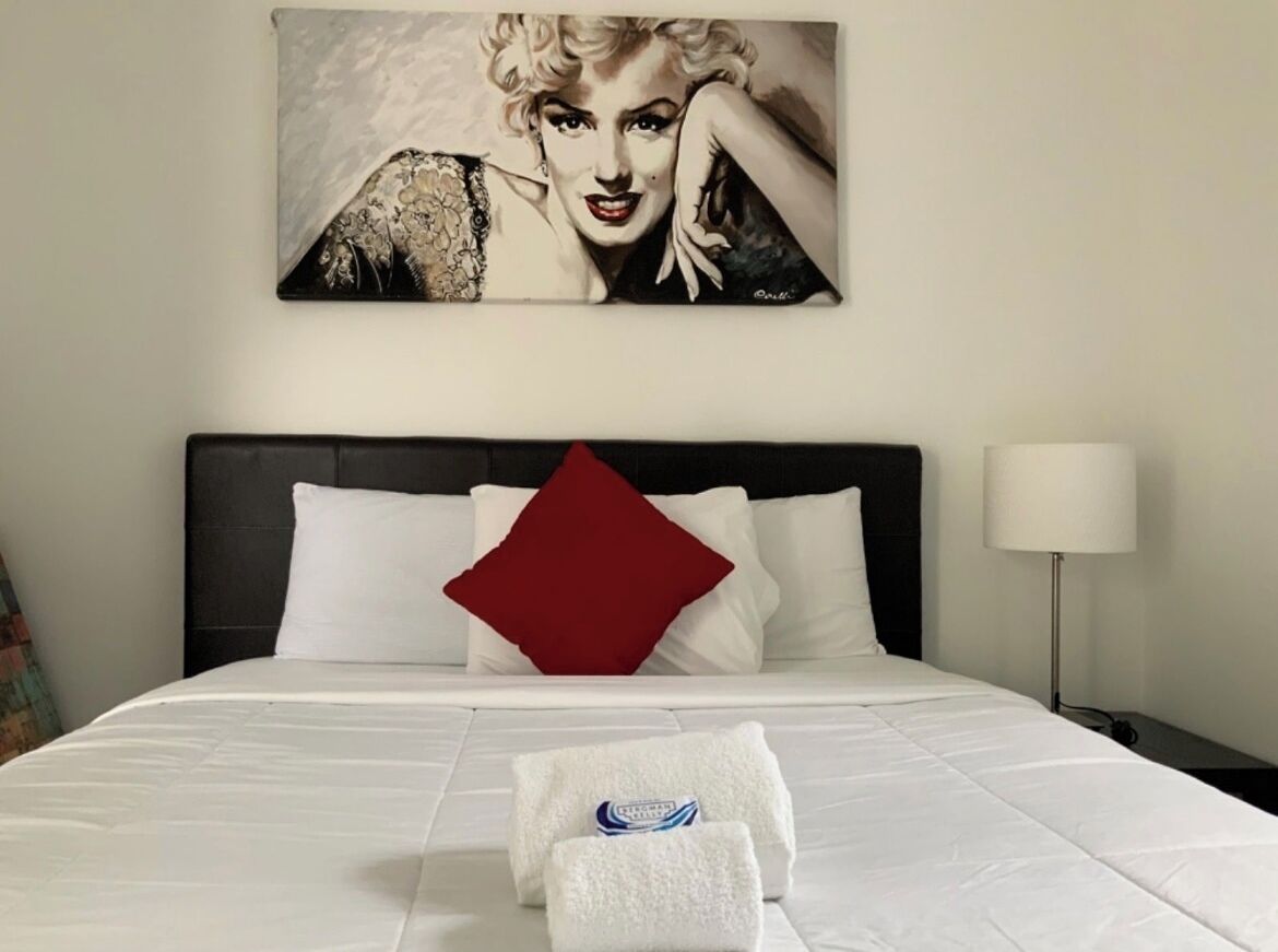 A neatly made bed with white bedding, a red pillow, and a towel with a logo.