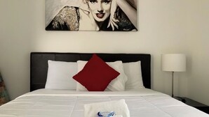 Una cama bien hecha con ropa blanca, un almohadón rojo y una toalla con un logotipo.
