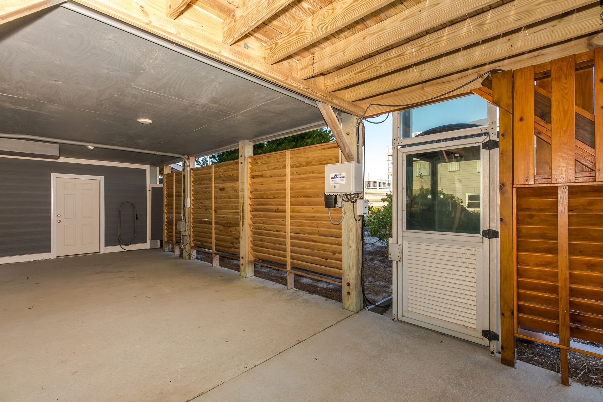 A covered outdoor area with a concrete floor, wooden beams, and a door leading to a residential building.