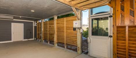 A covered outdoor area with a concrete floor, wooden beams, and a door leading to a residential building.