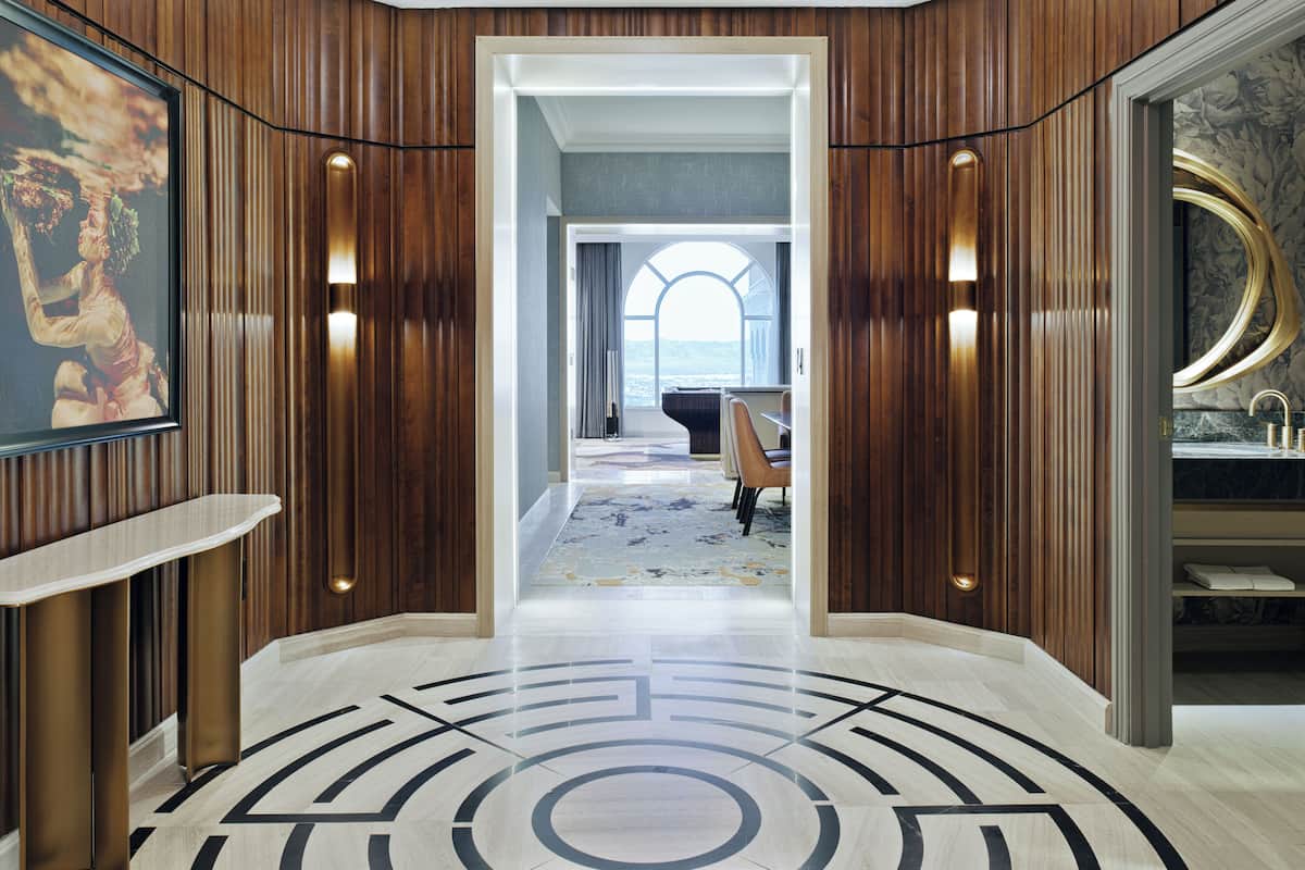 Un hall d’entrée luxueux avec des boiseries, un sol en marbre orné de motifs géométriques, un piano à queue et une vue sur la mer par une grande fenêtre en arc de cercle.