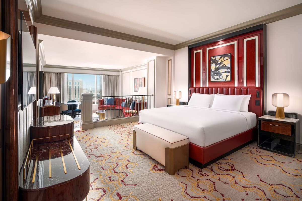 Une chambre d’hôtel dotée d’un grand lit, d’un coin salon avec un fauteuil et une table, d’un bureau et d’une grande fenêtre donnant sur la ville.