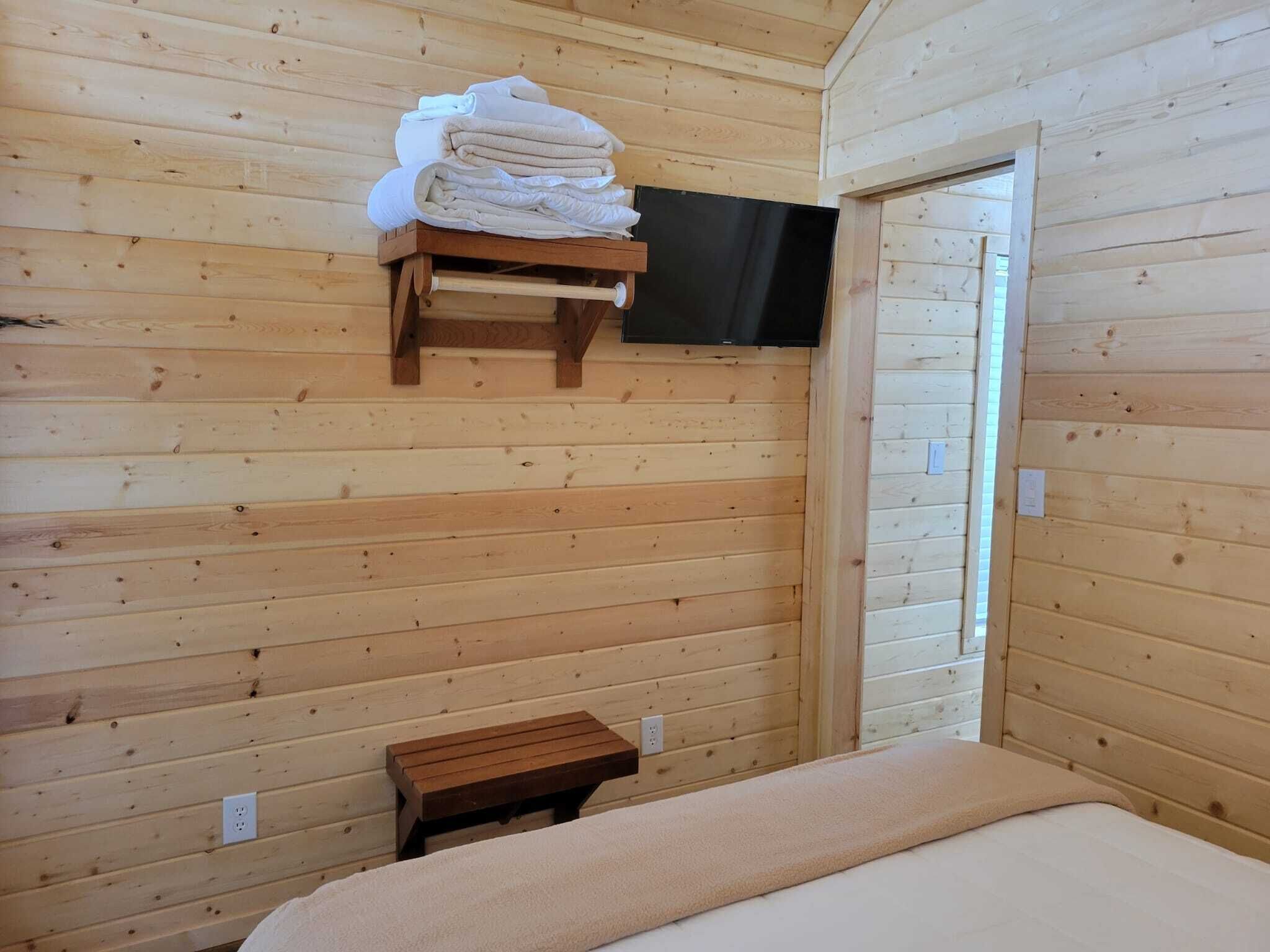 A wooden cabin bedroom with a bed, a small wooden bench, a shelf with towels, and a television mounted on the wall.