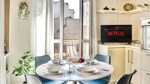 Un coin repas avec une table ronde dressée pour quatre personnes, un téléviseur affichant le logo de Netflix et une vue sur le paysage urbain par la fenêtre.