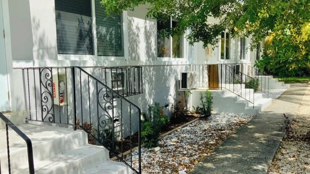 A residential building with a white exterior, a front entrance with a wooden door, and a small garden area with steps leading up to the entrance.