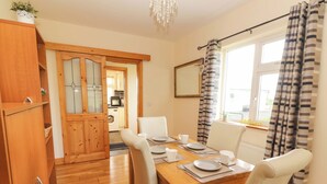 Un comedor con una mesa de madera dispuesta para cuatro personas, sillas blancas y vista a una cocina a través de una puerta abierta.