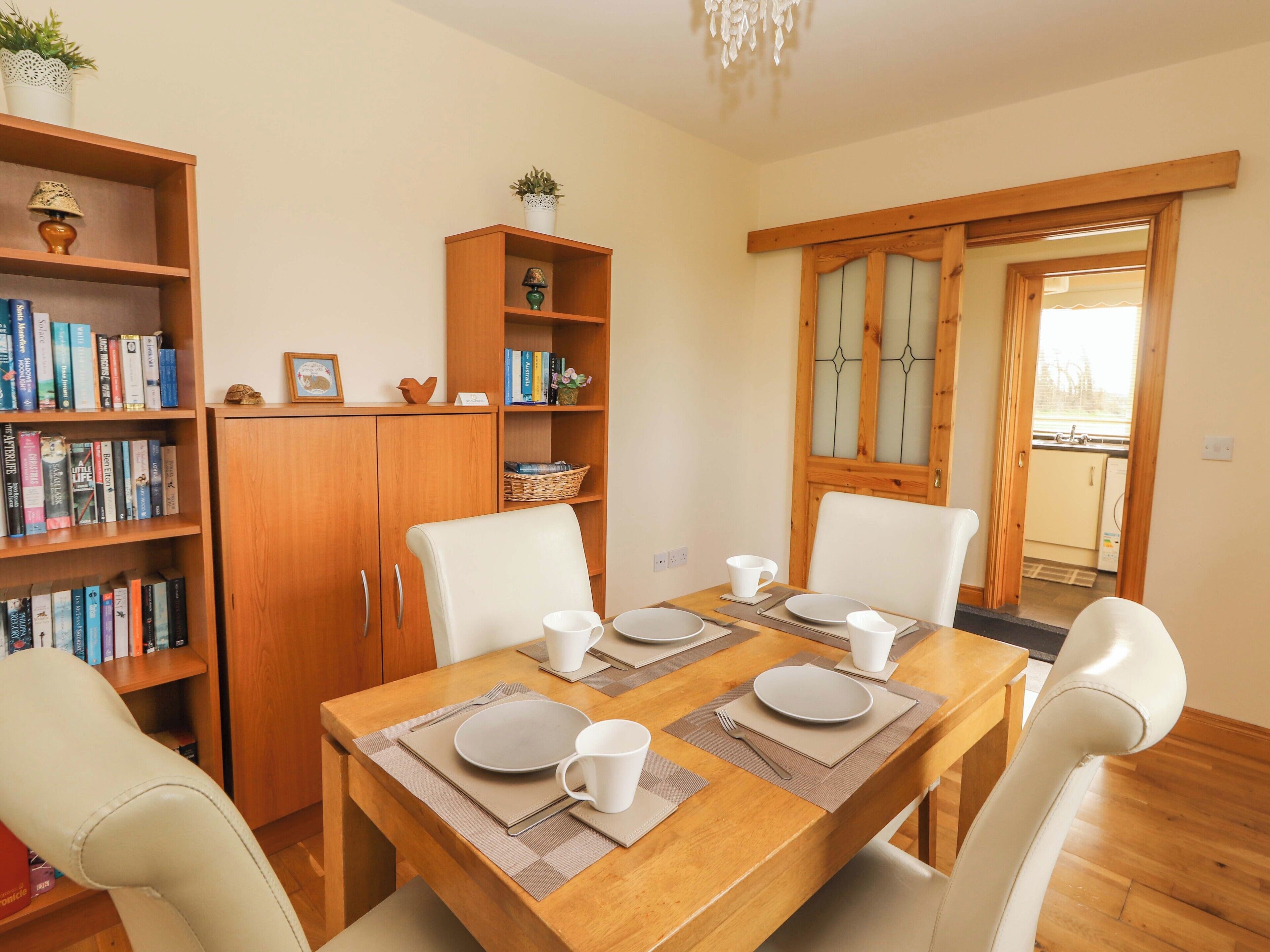 A dining area with a wooden table set for four, surrounded by white chairs. There are bookshelves on either side of the table, and a wooden cabinet is visible in the background.