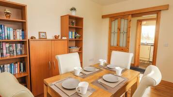 A dining area with a wooden table set for four, surrounded by white chairs. There are bookshelves on either side of the table, and a wooden cabinet is visible in the background.
