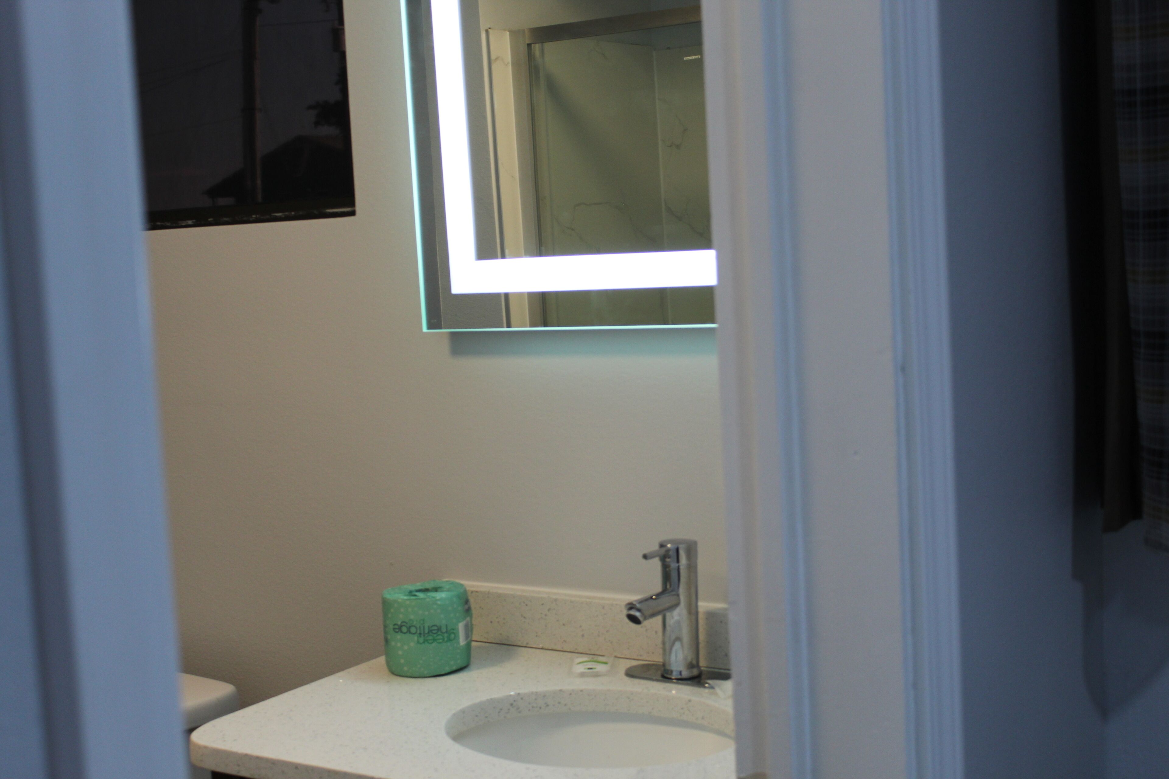 A bathroom with a white sink, a green soap dispenser, and a mirror with a light above it.