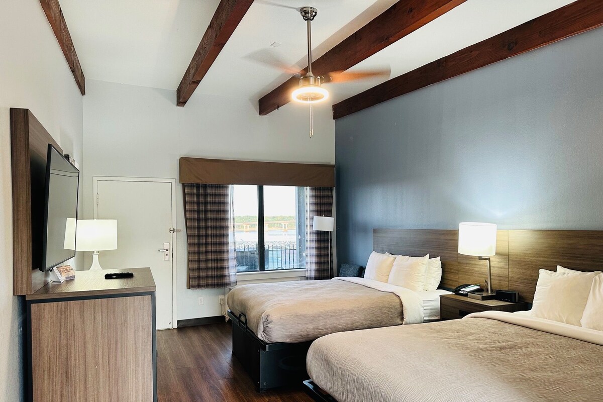 A hotel room with two beds, a desk, a mirror, and a ceiling fan.