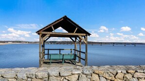 A wooden gazebo with a bench by a lake, a stone wall in the foreground, and a bridge in the background.