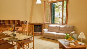 Una sala de estar acogedora con una mesa de vidrio, sillas de madera, un sofá color beige, una chimenea y un ventanal con contraventanas.