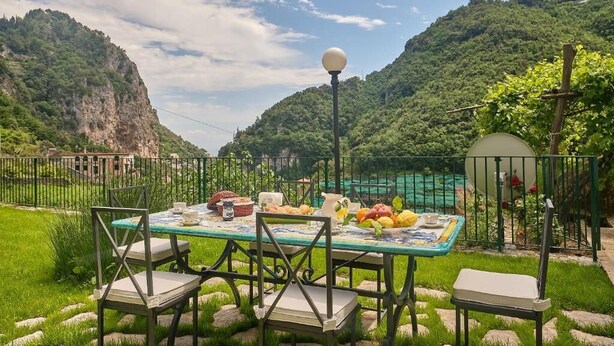 Mesa de comedor al aire libre con sillas, una lámpara y un mantel, frente a un paisaje montañoso.