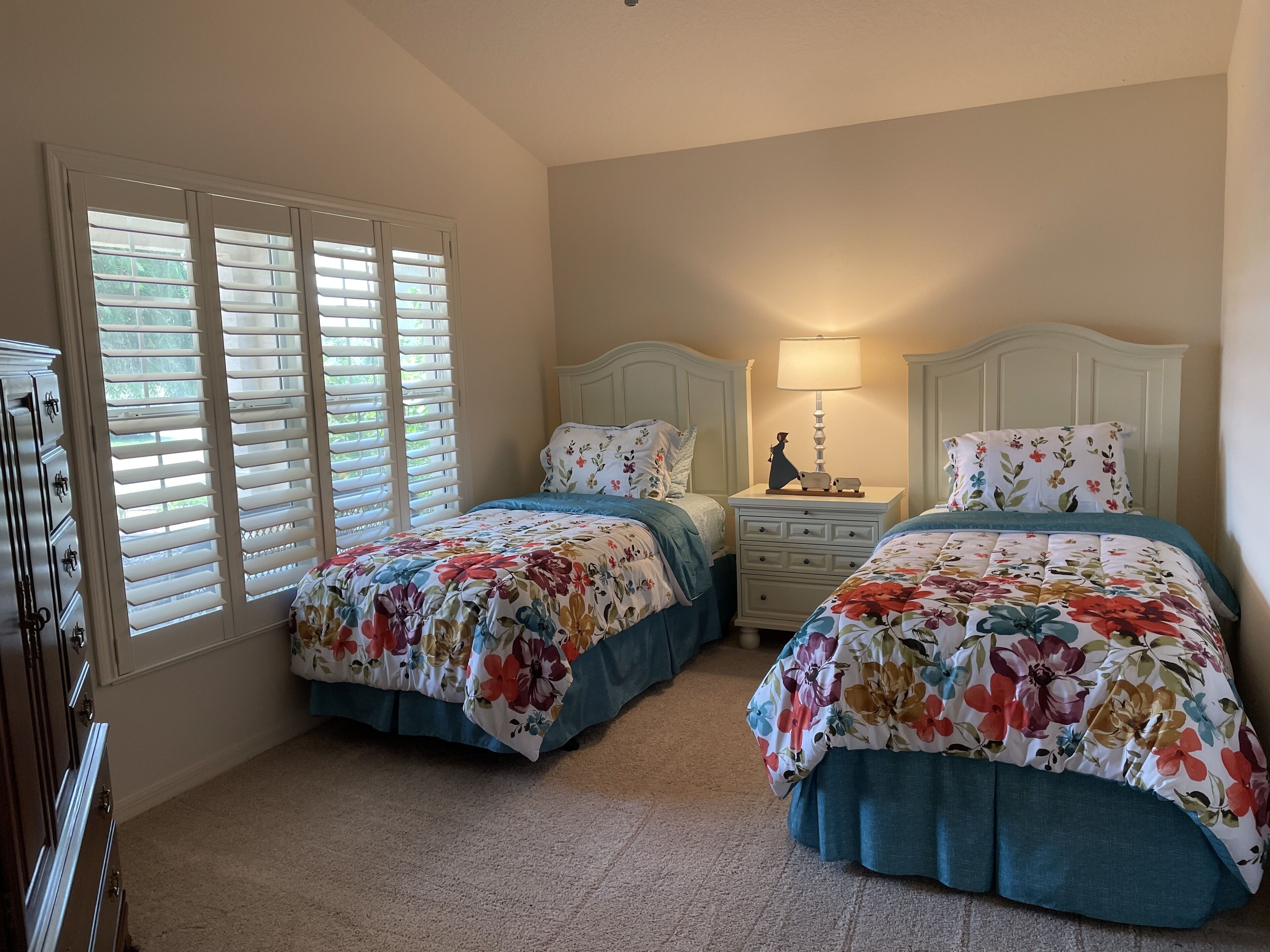 A bedroom with two beds, a dresser, a nightstand with a lamp, and a window with blinds.