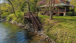 A cabin by a river with a wooden deck and stairs leading to a porch.