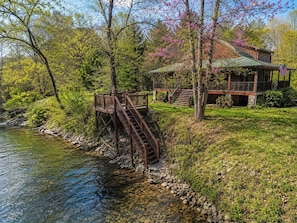 A cabin by a river with a wooden deck and stairs leading to a porch. - River Mornings, Hot tub Evenings, Repeat (Suches)