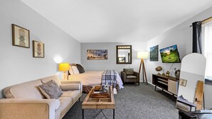 Ein Hotelzimmer mit Bett, Sofa, Couchtisch, Schreibtisch, Stuhl, Lampe, Fernseher und Bildern an den Wänden.