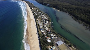 Vue aérienne d’une zone côtière comprenant une plage, une rivière et un quartier résidentiel avec des maisons et des immeubles.