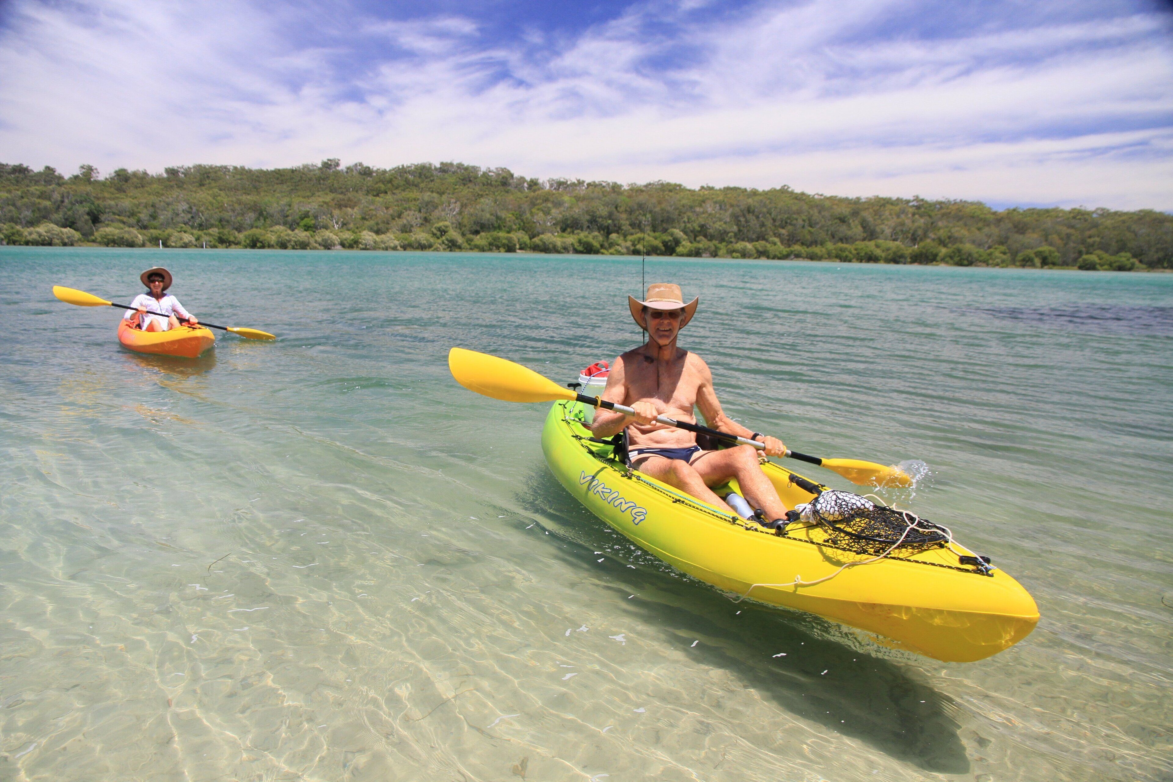 Two people kayaking on a calm, clear lake with a forested shoreline in the background.