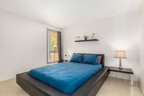 Relax in a serene bedroom with a large bed, minimalist decor, and a window with a curtain.