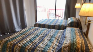 A hotel room with two beds, each with a patterned quilt. There is a window with curtains, a lamp on the wall, and a view of a balcony outside.